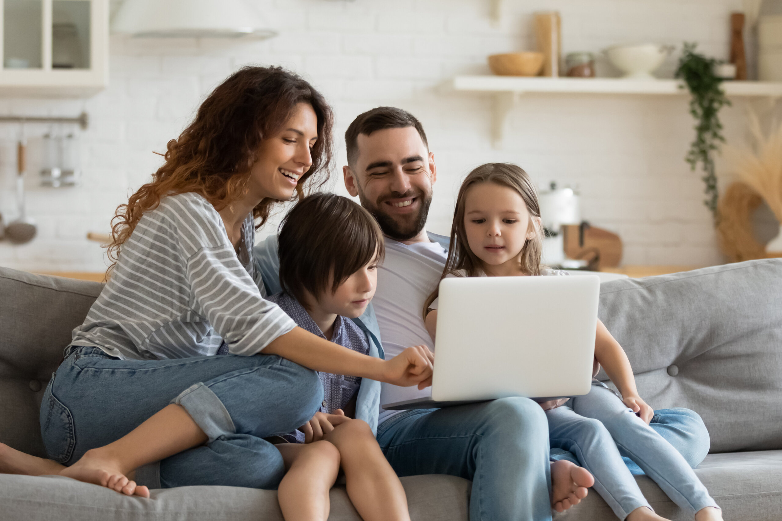 Happy young family with little kids sit on sofa in kitchen have fun using modern laptop together, smiling parents rest on couch enjoy weekend with small children laugh watch video on computer at home